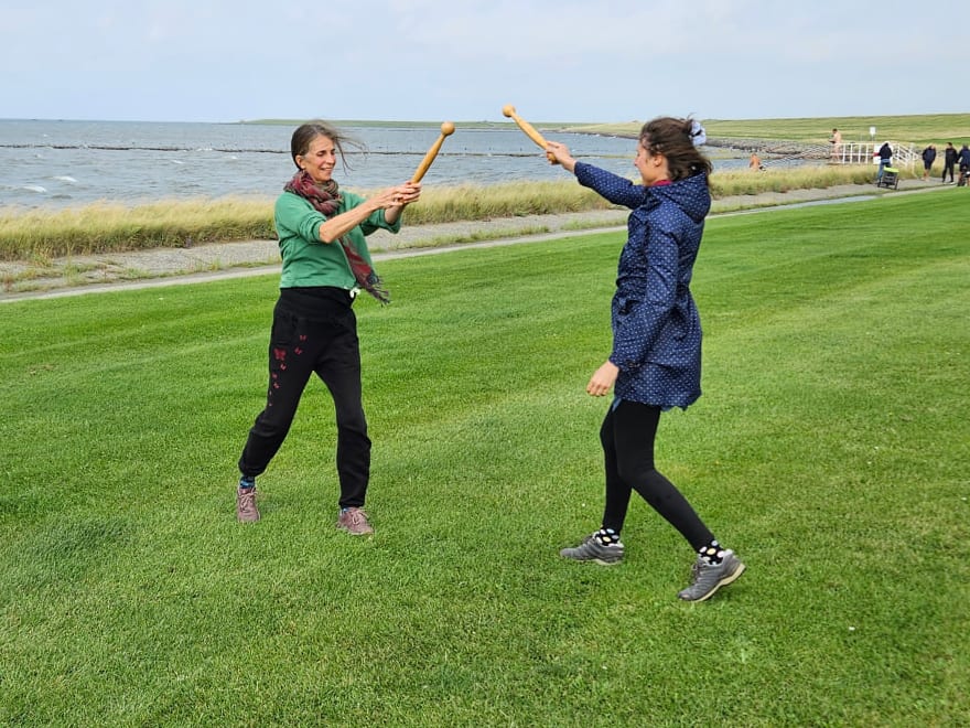 Sylke und eine junge Frau stehen sich auf einer Wiese am Meer gegenüber und führen ihre Idogostäbe zueinander