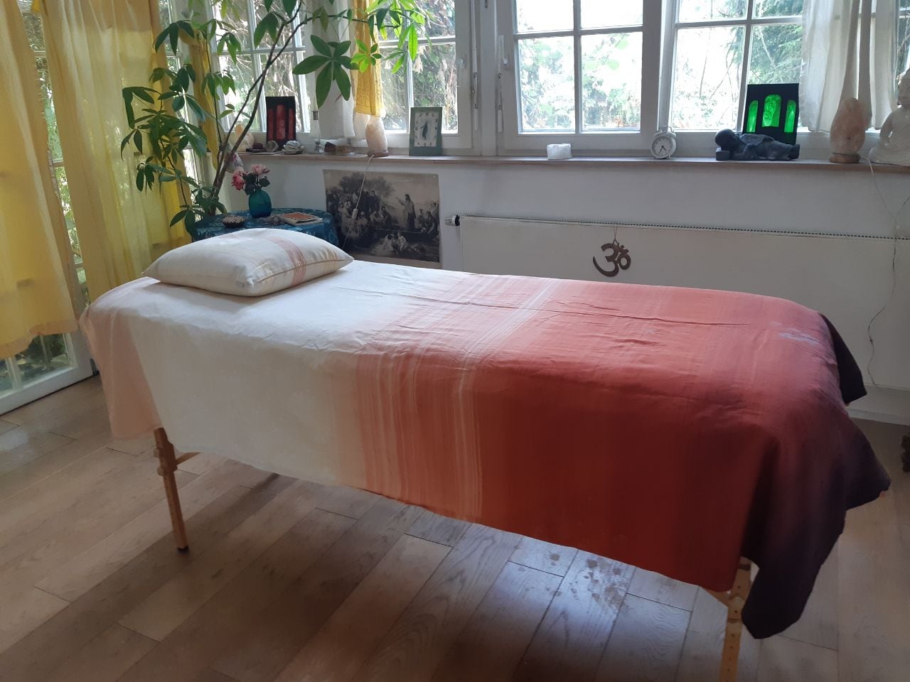 Behandlungsraum mit einer Liege unter hellen Fenstern, Pflanzen und gelben Vorhängen