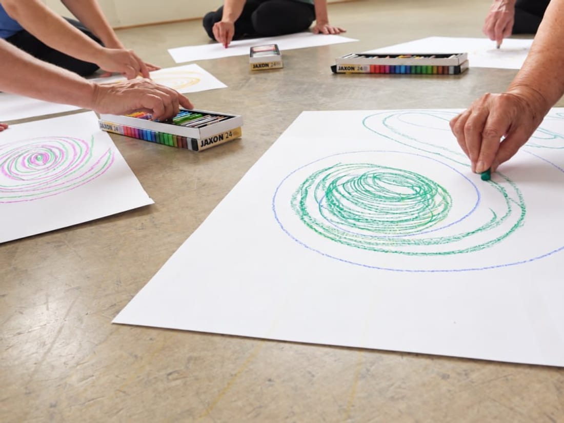 Mehrere Menschen knien am Boden und zeichnen mit Ölkreiden große Spiralen auf weiße Papierbögen
