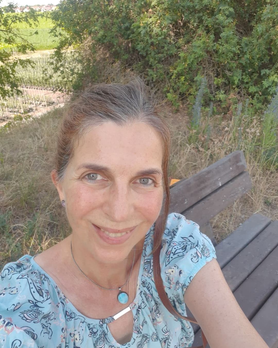 Sylke sitzt lächelnd auf einer Bank am Feldrand, im Hintergrund Weinberge