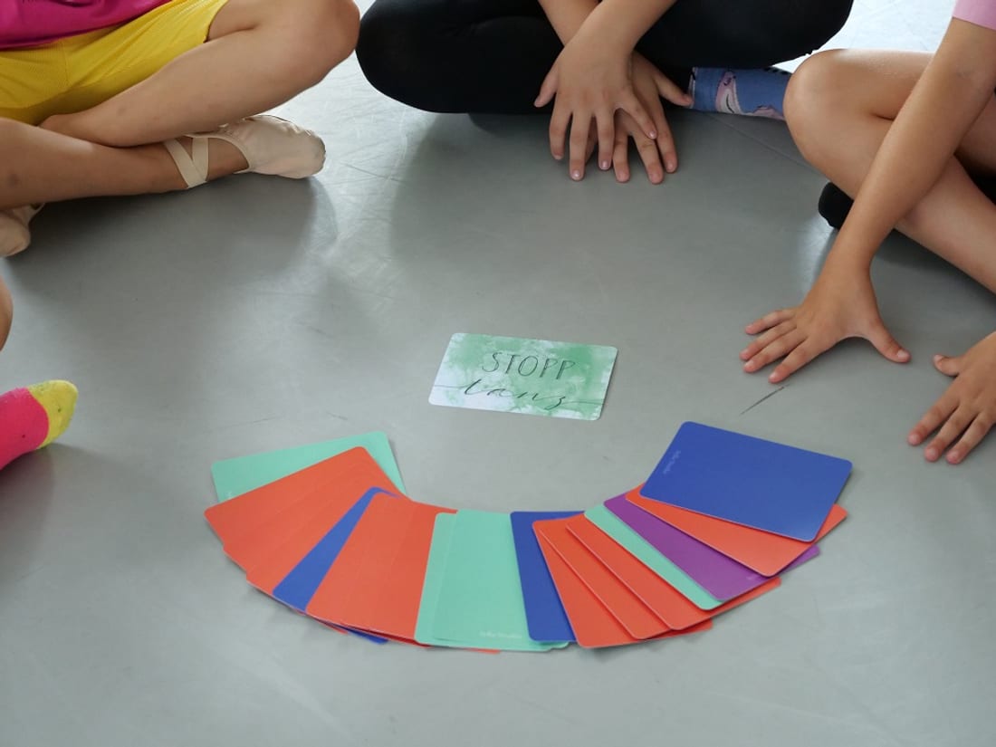 Kinder sitzen im Kreis um aufgefächerte bunte TanzSpielKarten und eine Karte mit der Aufschrift „Stopp tanz“