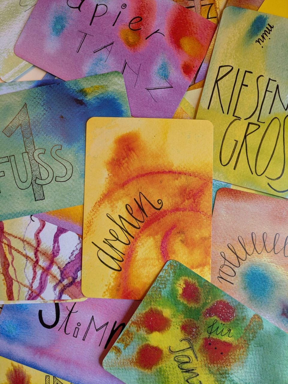 Bunt aquarellierte TanzSpielKarten mit kalligraphierten Worten wie „drehen“ und „Fuß“, lose übereinandergelegt