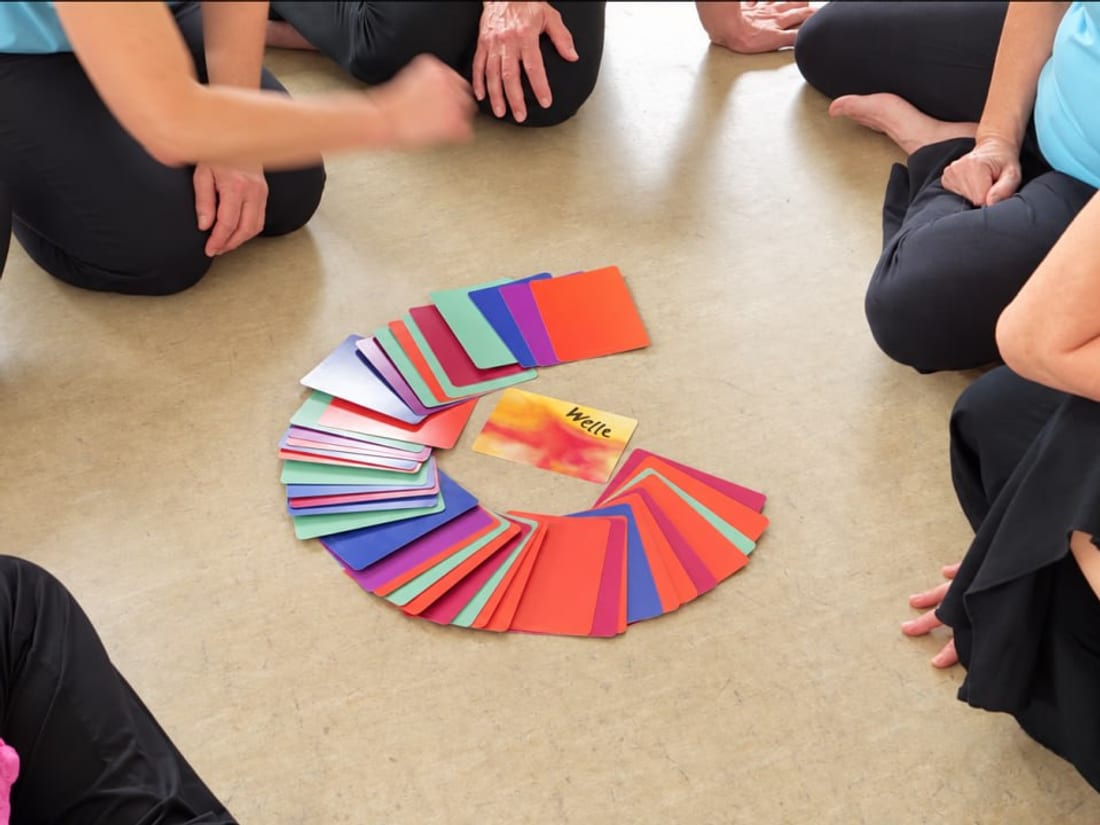Eine Gruppe sitzt am Boden um die bunt aufgefächerten TanzSpielKarten, in der Mitte liegt die Karte „Welle“ offen