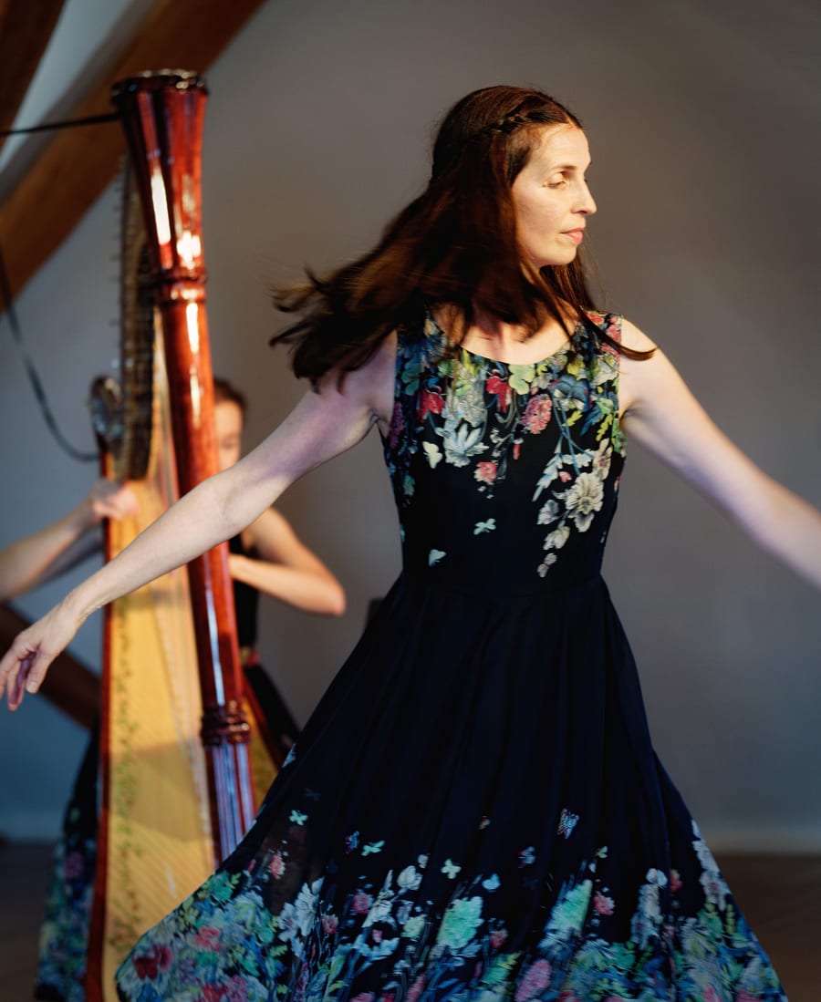 Sylke tanzt in einem geblümten Kleid mit ausgebreiteten Armen, hinter ihr spielt eine Harfenistin